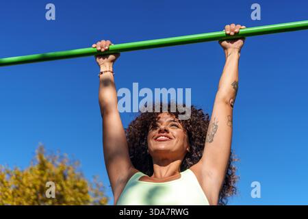 Woman hanging from a horizontal bar, smiling and looking up, engaged in a calisthenics workout for physical strength outdoors Stock Photo