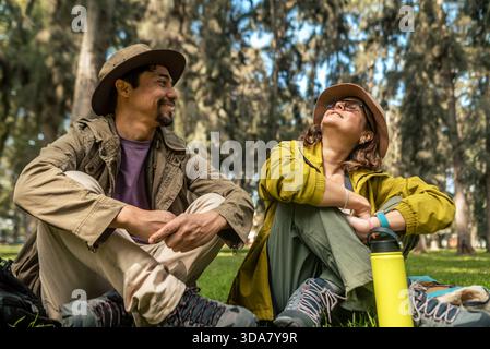 A man and a woman are sitting on the grass in a park. The man is wearing a hat and the woman is wearing a yellow jacket. They are smiling and seem to Stock Photo