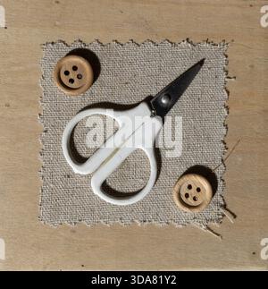Top view of scissors rest on a piece of fabric with two wooden buttons placed nearby. This setup is found on a wooden table. Stock Photo