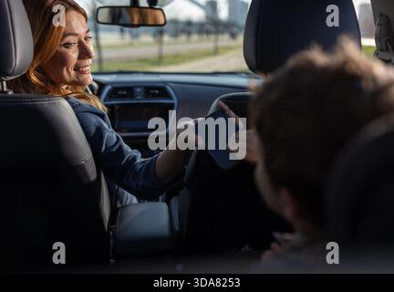 A moter sits in the front seat and smiles while handing a phone to a child in the back seat. They are enjoying their time on a road trip. Stock Photo