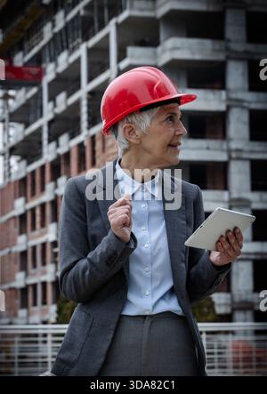 Architect in a hard hat looks at a tablet while standing near a building under construction. The site is active with workers and equipment around. Stock Photo