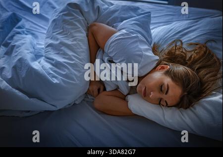 A woman rests in a bedroom at night, covered with a blanket, sleeping in a side position on her bed. Stock Photo