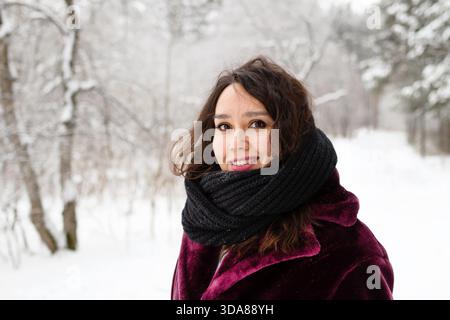 Beautiful smiling woman with long brown hair in a coat from faux fur and big black scarf on a background of winter forest. || Model released Stock Photo