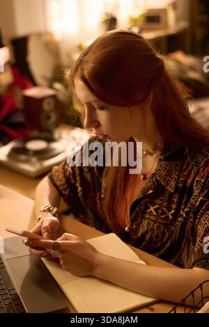 Caucasian young adult woman sitting at desk using smartphone while writing in notebook, concentrating on multitasking with laptop and stationery visible in workspace Stock Photo