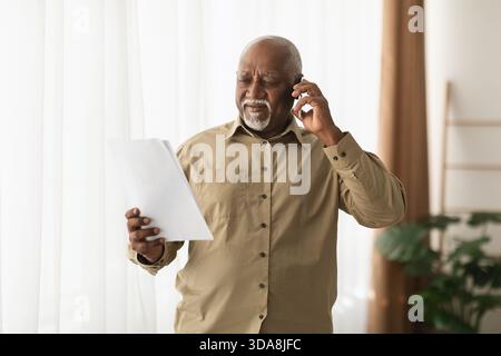Senior Man Engaged in a Phone Conversation While Reading Important Documents in a Well-Lit Living Space Stock Photo