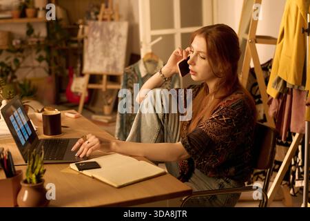 Young adult Caucasian woman sitting at desk working on laptop, holding phone to ear, surrounded by notebook and coffee cup, appearing focused in creative workspace Stock Photo