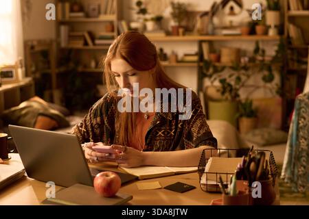 Young adult Caucasian woman sitting at desk using smartphone while working on laptop, surrounded by stationery and plants, focusing on multitasking in home office environment Stock Photo