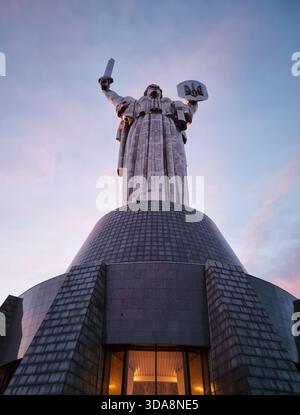 Mother Ukraine monument at sunset. Huge tall shiny metal statue of woman holding a sword and a shield against purple sunset sky with few clouds. Stock Photo