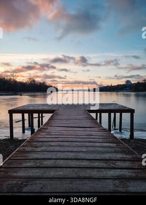 Wooden pier on the lake (symmetric view). Winter sunrise scene with ice over the water, trees and sky reflecting in mirror-like lake surface. Stock Photo