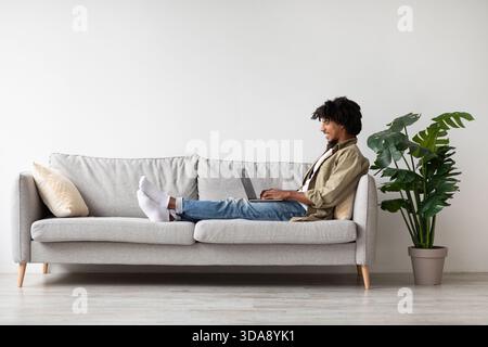 Cheerful Young Black Man Working From Home on Laptop While Relaxing on Couch Stock Photo