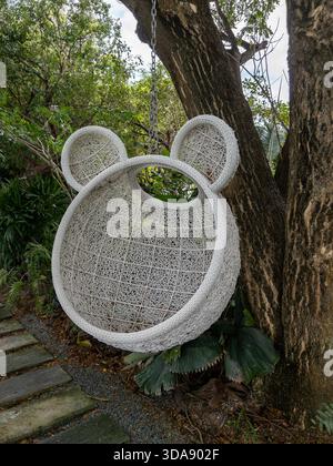 White rattan or wicker hanging basket chair, shaped like an animal head, suspended from a tree in a lush outdoor coffee shop garden. Stock Photo