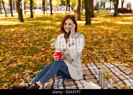 Smiling Woman Enjoys a Cozy Moment in a Park Surrounded by Autumn Leaves While Sipping From a Red Cup Stock Photo