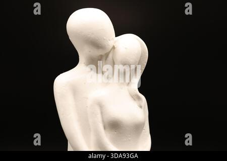 Minimalist white sculpture of two abstract human figures embracing against a dark background. Stock Photo