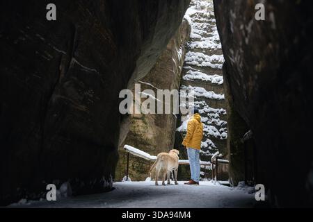 Man walks with dog along narrow snowy path between high rocks. Labrador retriever stays close to owner in cold winter landscape. Stock Photo