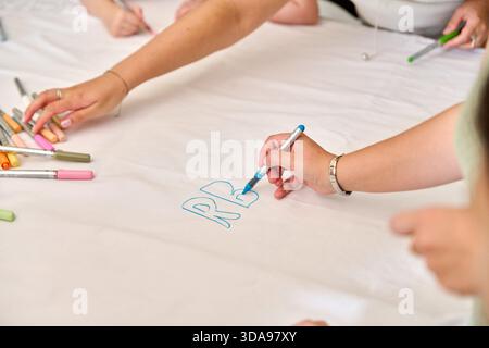 Multiple hands actively drawing and coloring on large white paper surface using various colored markers. Bright lighting highlights creative collabora Stock Photo