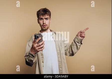 Young man with surprised expression holding smartphone and pointing. Stock Photo