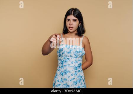 Young woman in floral dress pointing at camera against beige background. Stock Photo