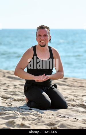 Smiling adult man with short hair sits in kneeling yoga pose on sandy beach by calm Baltic Sea water. Bright natural light enhances serene and mindful Stock Photo