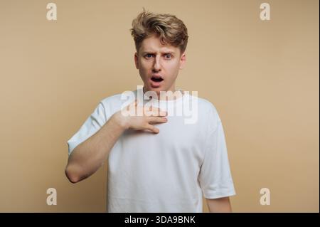 Young man in white t-shirt looking surprised against beige background. Stock Photo