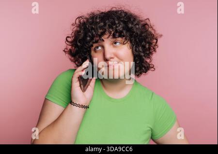 A young woman with curly hair is casually talking on her smartphone. She wears a bright green shirt and has a playful expression while standing in fro Stock Photo