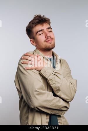 A young man stands in a light studio, wearing a relaxed outfit and gently hugging himself with a serene expression, promoting a feeling of self-accept Stock Photo