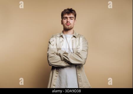 A young man stands with his arms crossed, showing a relaxed yet confident demeanor. The neutral background highlights his casual style and expression, Stock Photo