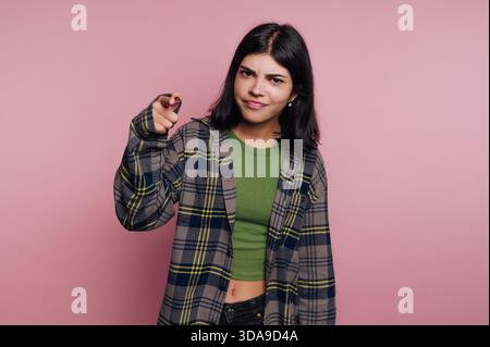 A young woman stands confidently against a pink background, wearing a green top and a plaid shirt. She playfully points her finger as if to emphasize Stock Photo