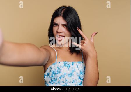 A young woman poses for a selfie, smiling and making a fun gesture with her hands. She stands against a simple beige background, showcasing her playfu Stock Photo