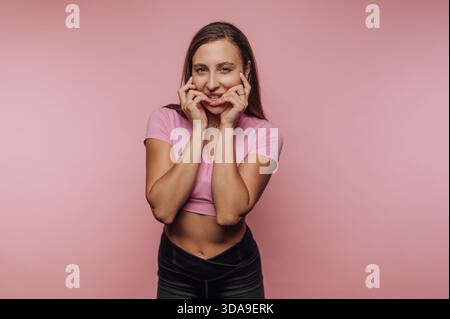 A young woman stands confidently in front of a vibrant pink backdrop. She smiles wide and poses playfully, wearing a fitted pink top and black pants. Stock Photo