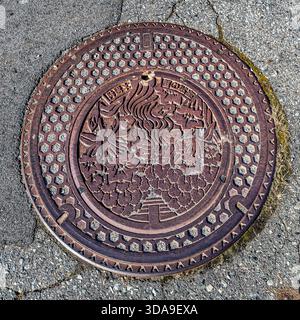 Cast-iron manhole cover with a decorative relief from the Kiso Valley – stylised plant motifs and a bird figure on a dark, slightly moss-covered background. In Japan, each local authority designs its own manhole covers as a symbol of local identity. Kiso, Nagano Prefecture, Japan Stock Photo