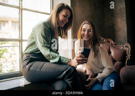 Two female colleagues sharing a relaxed moment while looking at a smartphone in a bright modern office, representing teamwork, friendship and positive Stock Photo
