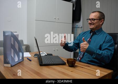 A cheerful mature businessman in a teal shirt is sitting at his wooden desk giving two thumbs up during an online video conference Stock Photo