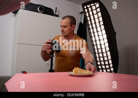 A focused mature man in a mustard t-shirt is standing and adjusting a camera on a tripod to photograph a cookie on a pink backdrop Stock Photo
