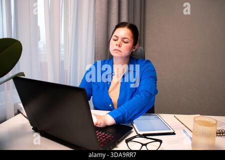 Concentrated woman with glasses types on laptop at home desk. Natural light from window highlights her productive remote work session. Focused young w Stock Photo