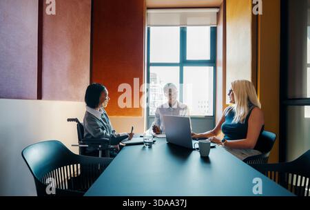 Three diverse women, one using a wheelchair, are engaged in a meeting, collaborating and sharing ideas. They sit at a long table with a laptop in fron Stock Photo