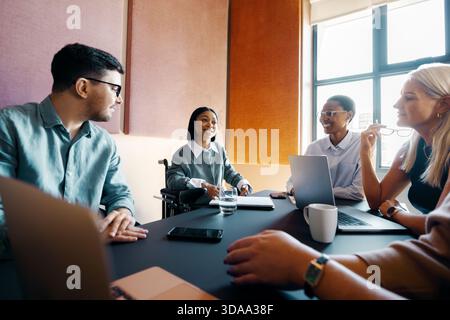 Colleagues, including a wheelchair user, engage in a collaborative meeting with laptops and documents. Stock Photo
