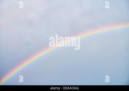 Delicate rainbow stretches gracefully across pale blue sky with subtle cloud formations, creating tranquil and uplifting natural scene Stock Photo