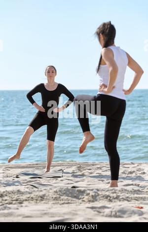 Adult woman yoga instructor wearing black performs balancing pose while coaching woman in white top on sandy Baltic Sea shore under clear daylight sky Stock Photo