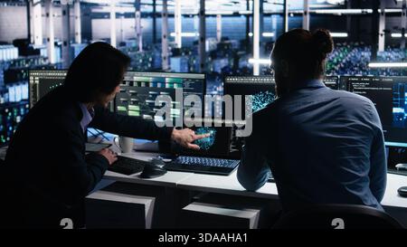 Data center system administrators use PC to monitor neural network using AI LLM. Asian man and colleague at work using artificial intelligence to oversee servers powering machine learning, camera A Stock Photo