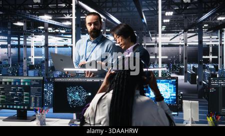 Team of engineers doing brainstorming in server room using AI to review infrastructure diagnostics. Teamworking IT colleagues in data center analyzing data using artificial intelligence, camera A Stock Photo