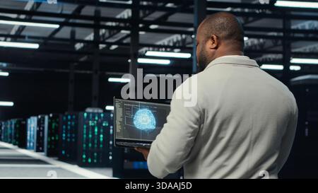 Technician using laptop in data center, managing server virtualization, running scripts. IT expert using programming language on notebook in server hub, finetuning network protocols, camera B Stock Photo
