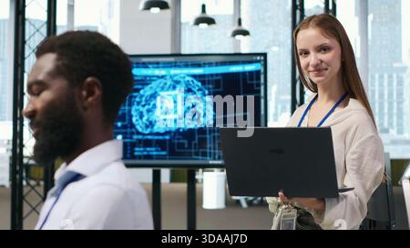 Portrait of smiling female engineer coding with AI language model assistance in startup office, using notebook. Happy young worker typing on laptop, using LLM assistant, camera A Stock Photo