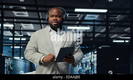 Data center admin wearing headphones using tablet to identify bottlenecks. Server farm IT expert listening music while troubleshooting bugs, implementing optimizations, camera B Stock Photo