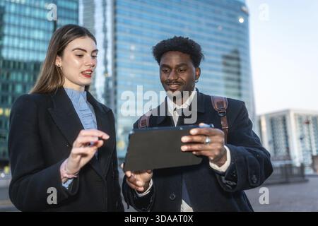 Two diverse business professionals collaborate outdoors in a modern city, smiling as they review data and strategy on a sleek tablet, sharing ideas an Stock Photo