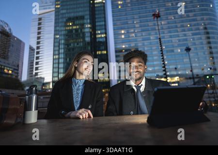 Diverse business colleagues working together on a digital tablet outdoors on a wooden table, collaborating in a city setting with illuminated modern s Stock Photo
