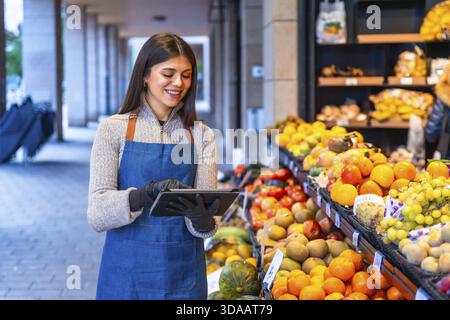 Smiling woman grocer in apron and gloves uses a tablet to manage inventory and orders at a fresh produce stand, modern small business efficiency and c Stock Photo