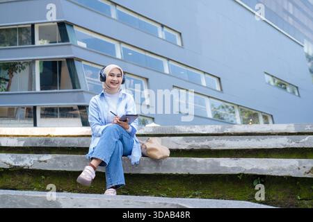 A young Asian woman wearing headphones and glasses holds a tablet while standing in a green park near campus buildings. She smiles confidently, repres Stock Photo