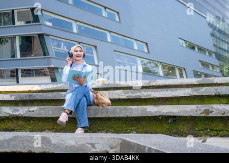 A young Asian woman wearing headphones and glasses holds a tablet while standing in a green park near campus buildings. She smiles confidently, repres Stock Photo