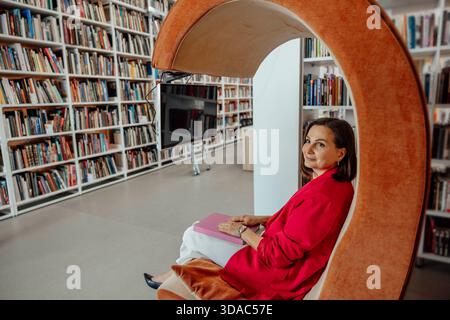 young Hispanic woman with brown hair sits in a modern orange chair, working on a laptop in a coworking space filled with bookshelves. Stock Photo