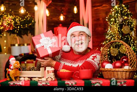 Joyful atmosphere. Senior joyful man Santa claus drinking tea. Winter holiday. Joyful grandpa with gift. Dreamy grandfather in Santa hat. Christmas Stock Photo